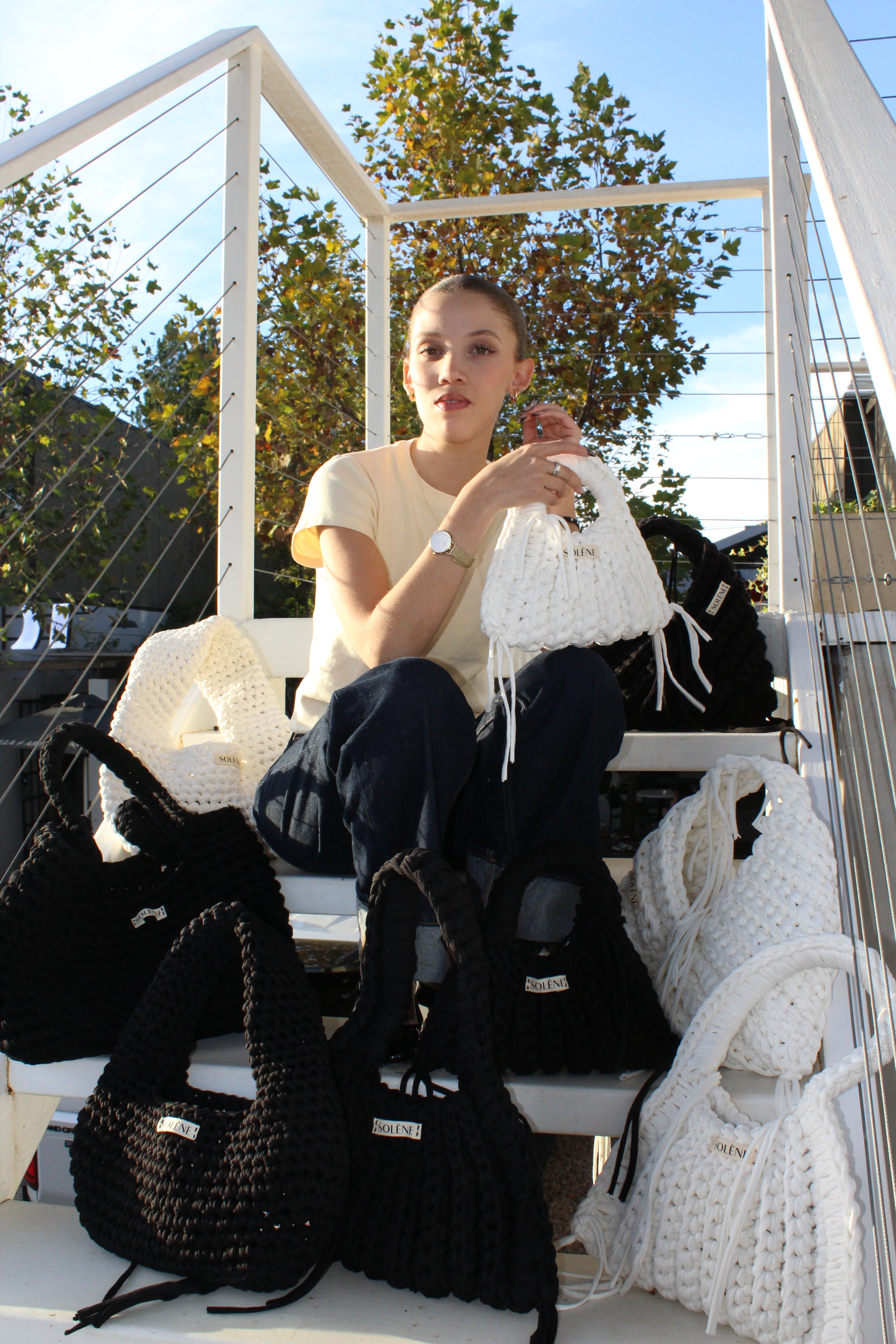 Woman sitting on a white chair with black bags around her, outdoors.